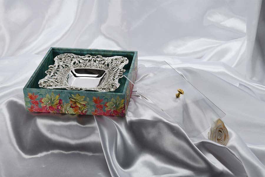 A square shaped silver plated bowl decorated with intricate patterns on the border giving it an antique look inside a magnificent gift box of pink, turquoise & blue color