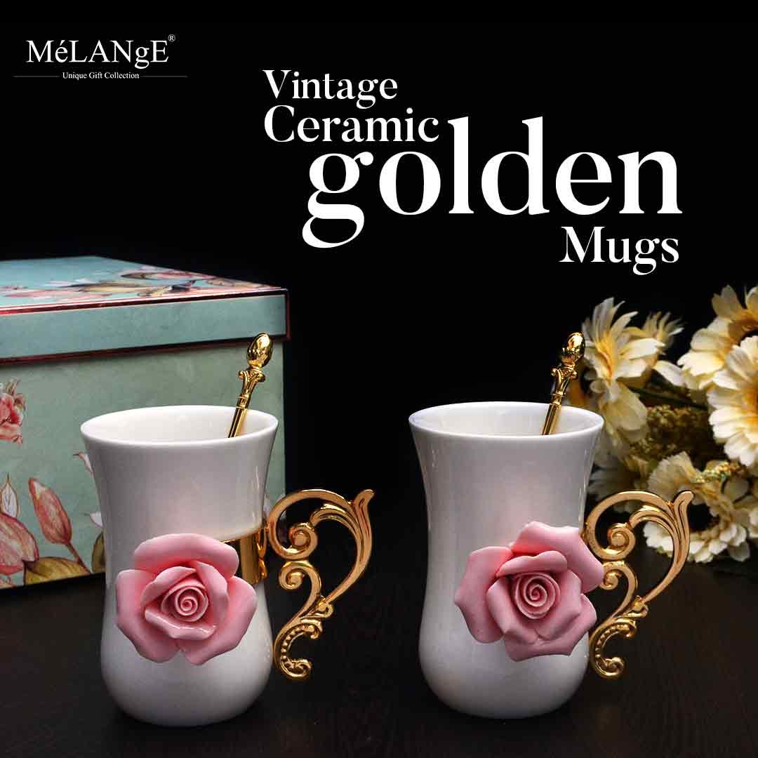 Vintage Ceramic Golden Mug with intricate roses embellished on their fronts along with golden spoons sit on a dark surface with flowers and a gift box in the background.